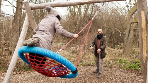 A child in a brightly coloured red and blue buddy swing, being pushed by a smiling man. It is winter.